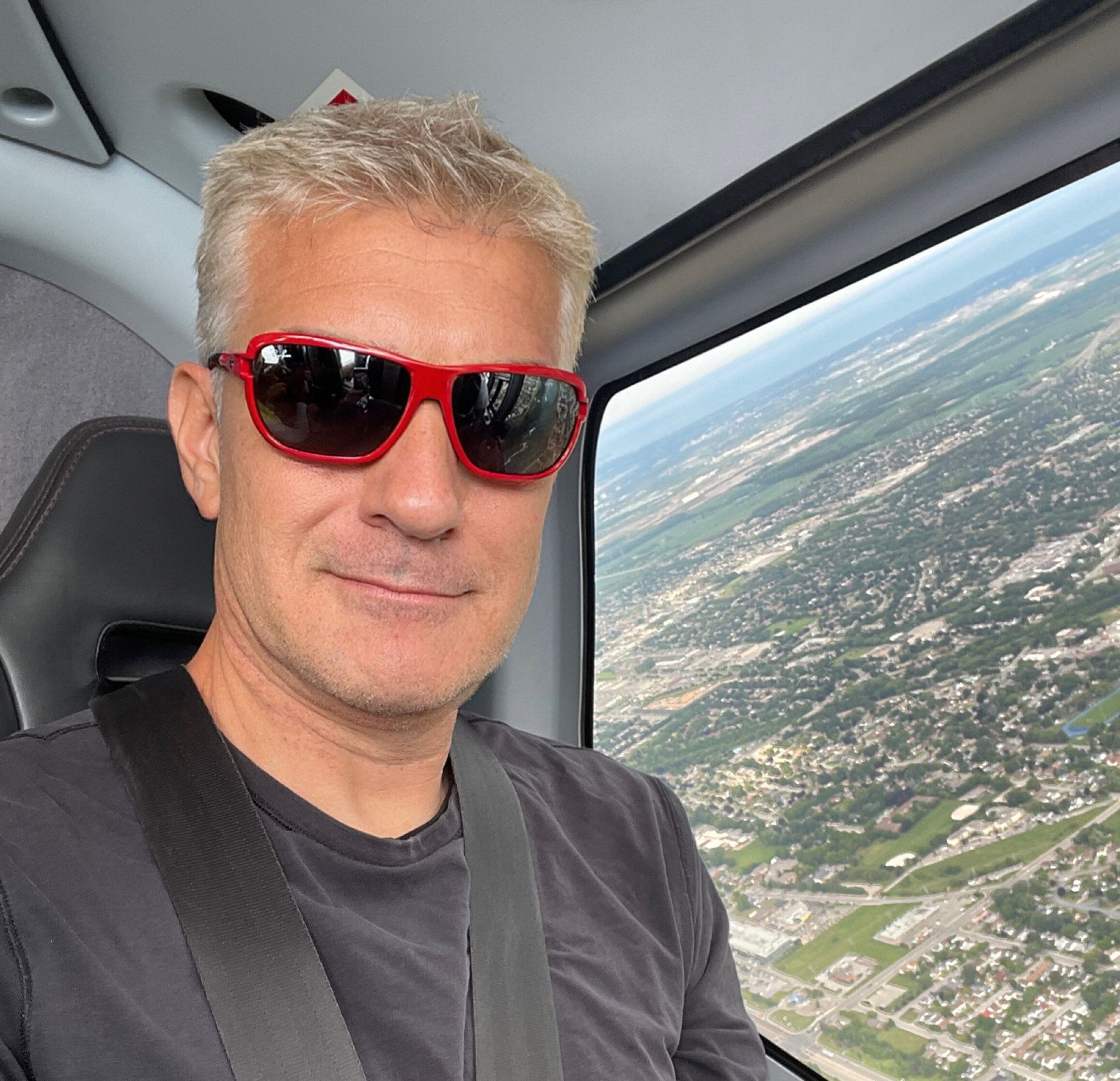 Sascha Ghods mit roter Sonnenbrille im Flugzeug, durch das Fenster die Stadtlandschaft tief unten.