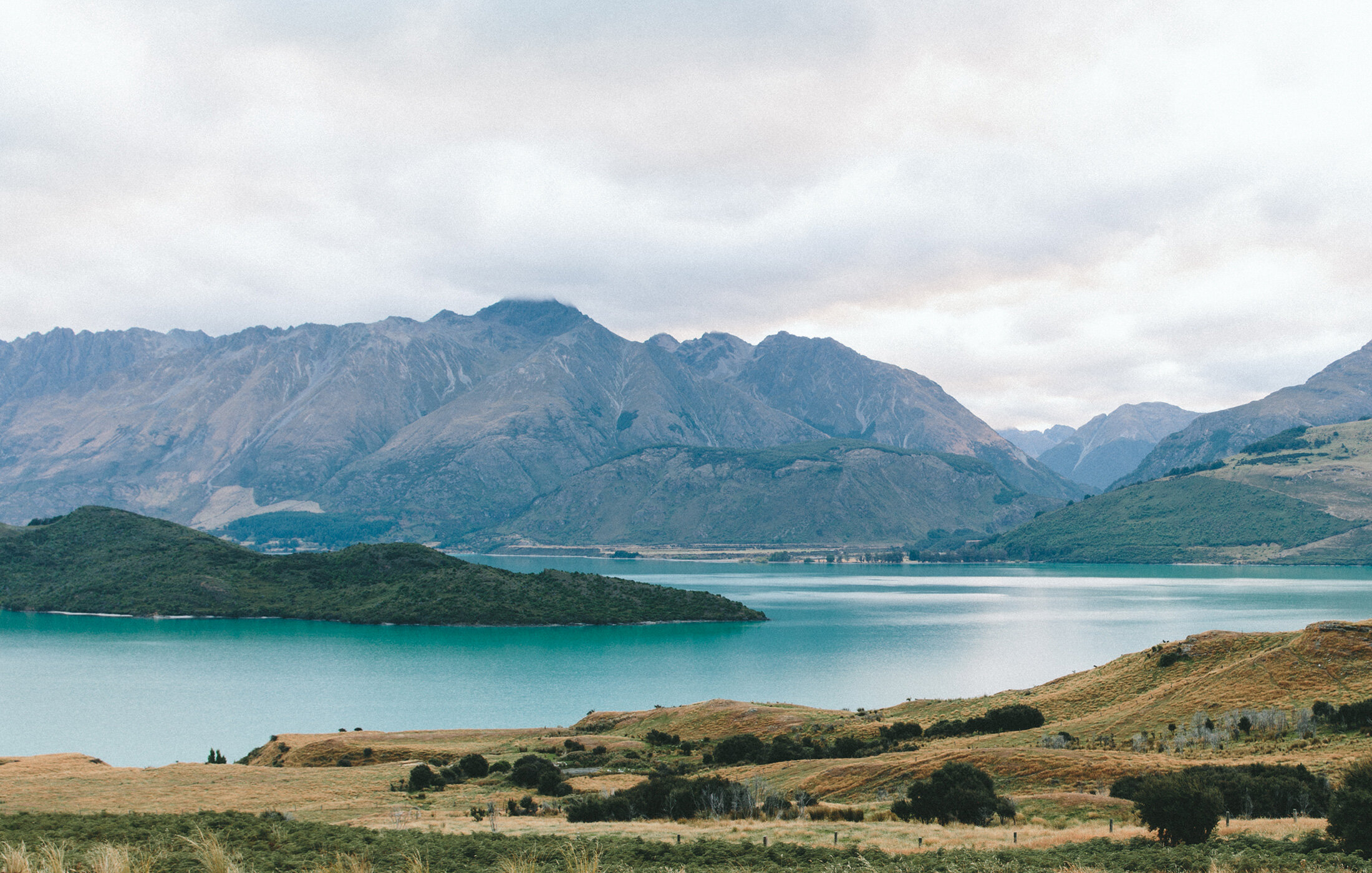 Scenic view of mountains and a lake with grassy hills in the foreground under a cloudy sky.