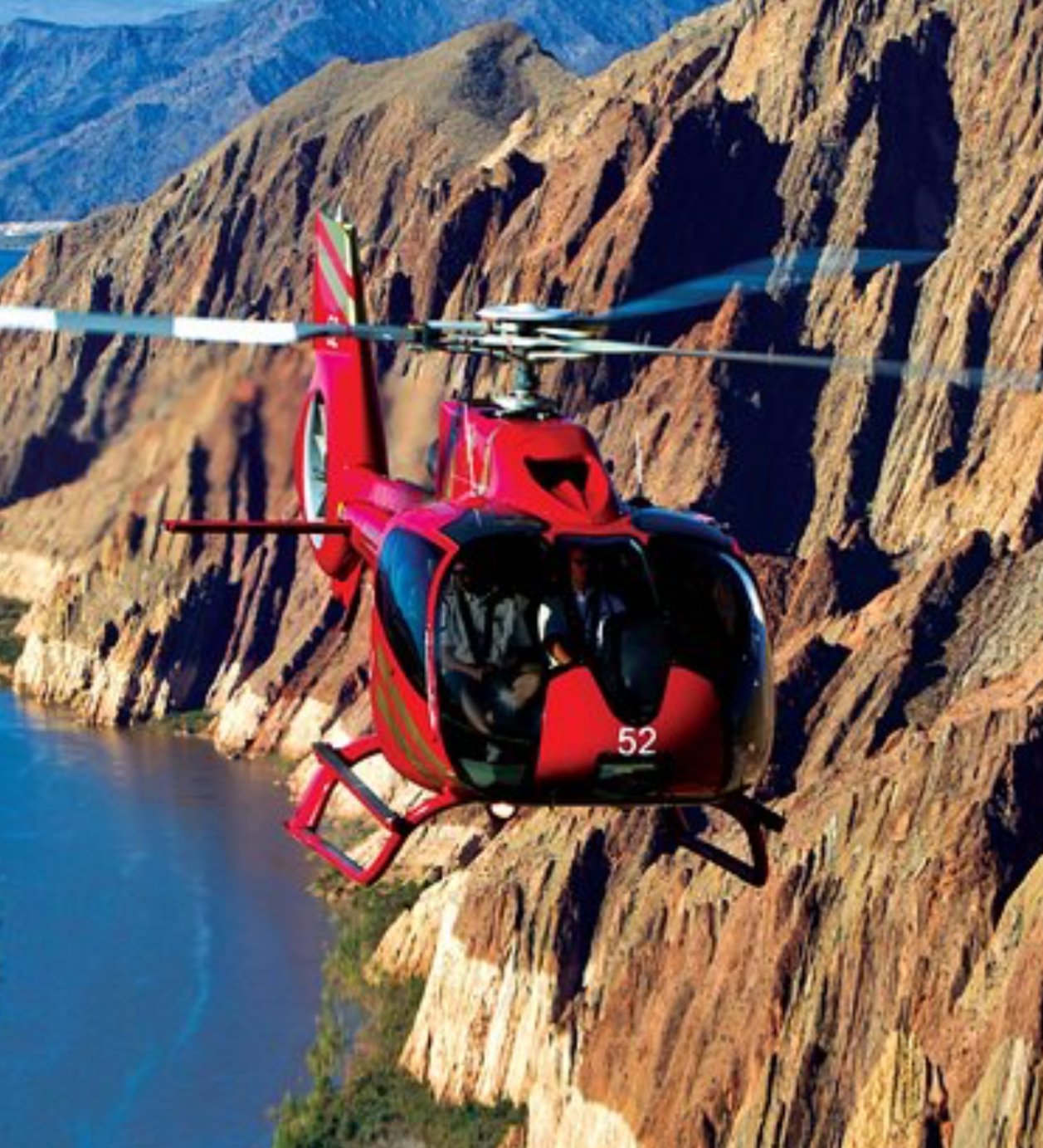 Red helicopter flying over the Grand Canyon with cliffs and river below.
