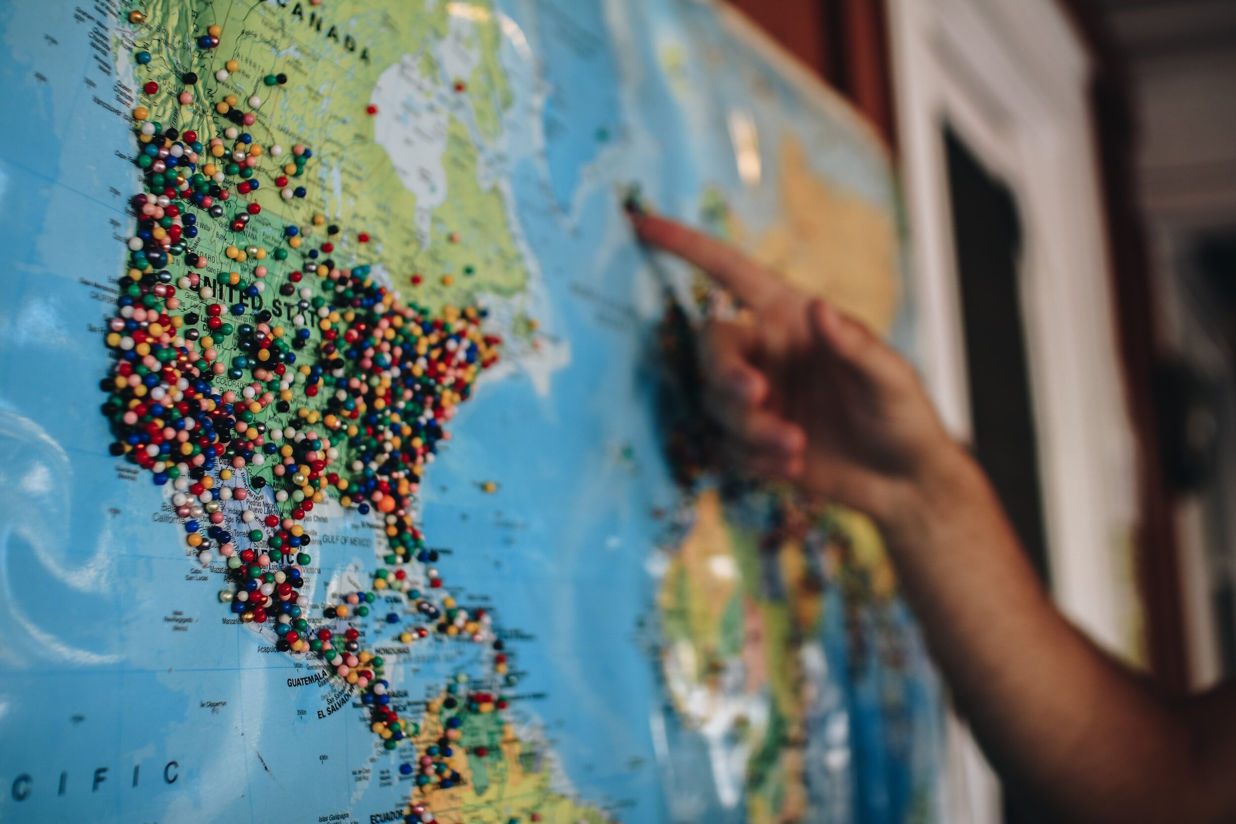 Person pointing at a world map covered with colored pins marking various locations.