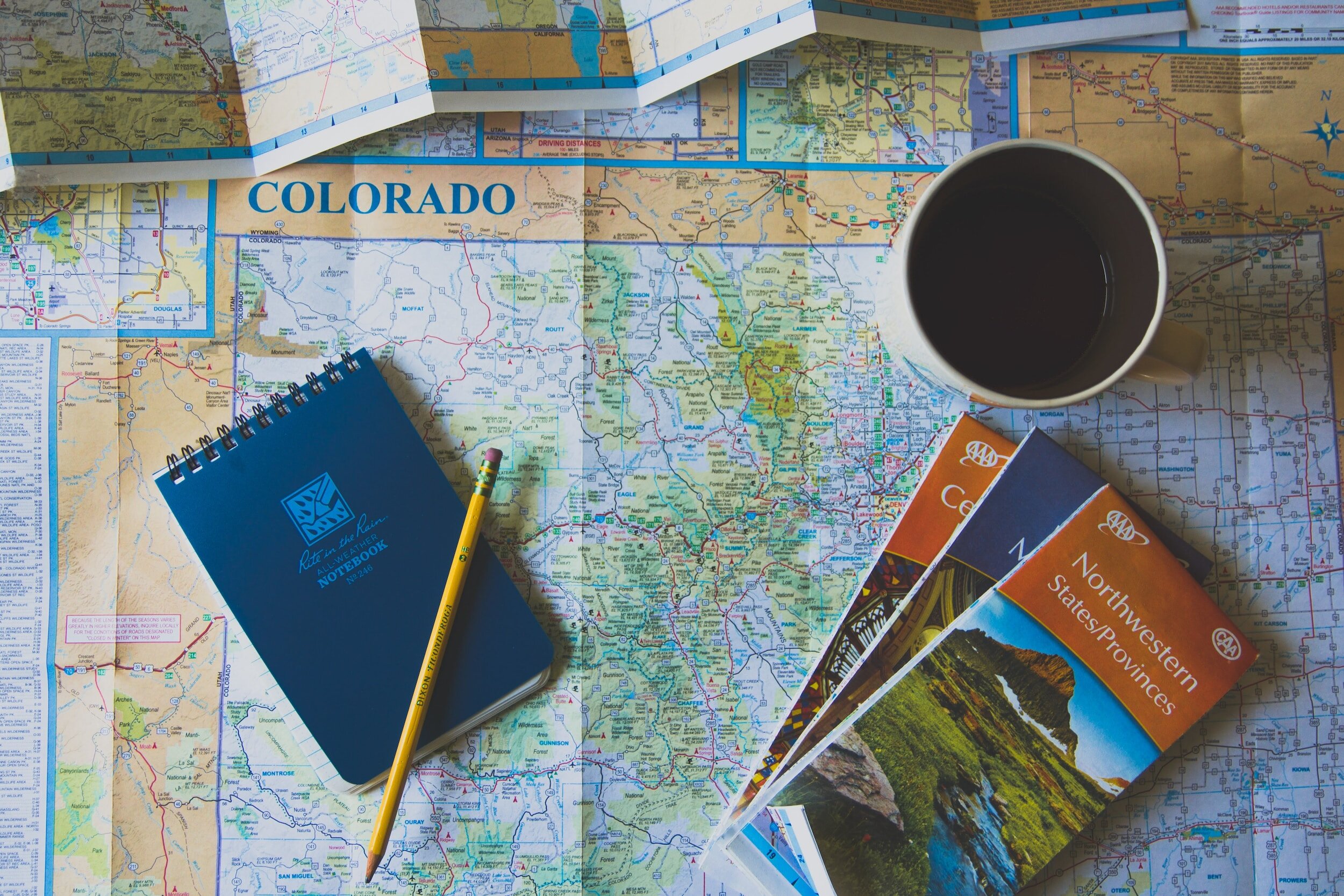 Travel planning items including a map of Colorado, a notebook with a pencil, a cup of coffee, and AAA travel brochures.